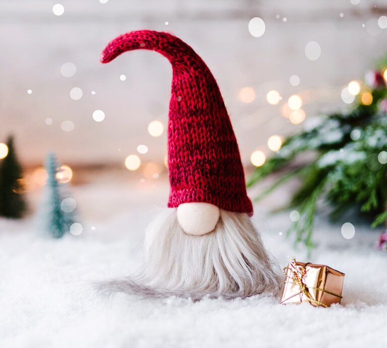 Ein selbst gebastelter Wichtel aus Socken mit roter Strickmütze und grauem Bart steht auf schneebedecktem Boden, daneben ein goldenes Geschenk. | © Adobe Stock/ emmi