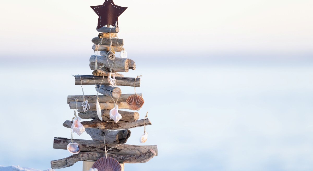 Aus Treibholz und Muscheln gebastelter Weihnachtsbaum mit Sternspitze steht auf hellem Sand vor sanftem Meereshorizont.