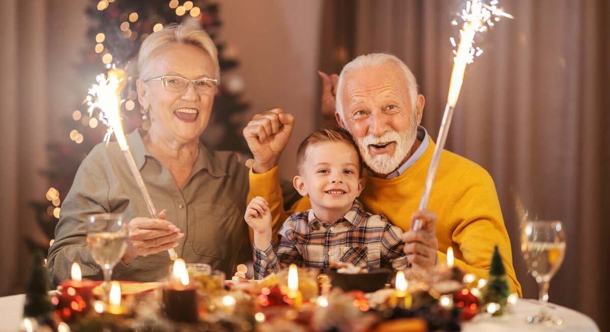 Älteres Paar und ein Junge sitzen an Silvester lächelnd mit Wunderkerzen an einem festlich gedeckten Tisch vor einem geschmückten Weihnachtsbaum.
