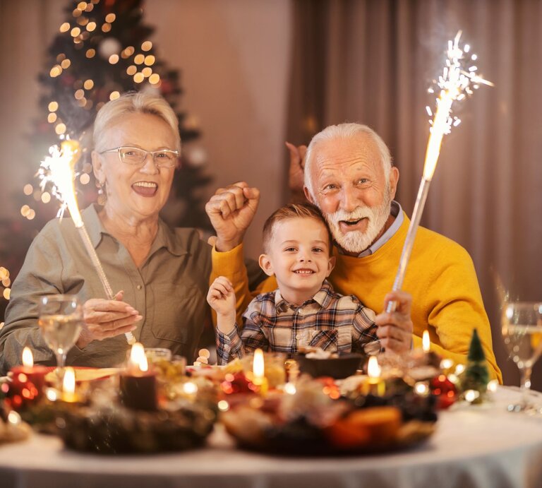 Älteres Paar und ein Junge sitzen an Silvester lächelnd mit Wunderkerzen an einem festlich gedeckten Tisch vor einem geschmückten Weihnachtsbaum. | © Adobe Stock/dusanpetkovic1