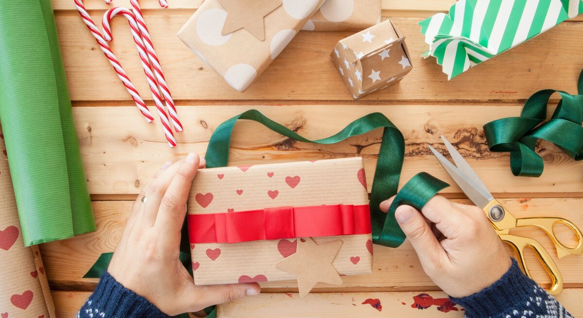 Zwei Hände in blauem Strickpulli verpacken ein Geschenk in Herzpapier mit rotem Band, umgeben von Geschenkpapier, Schleifen und Zuckerstangen.