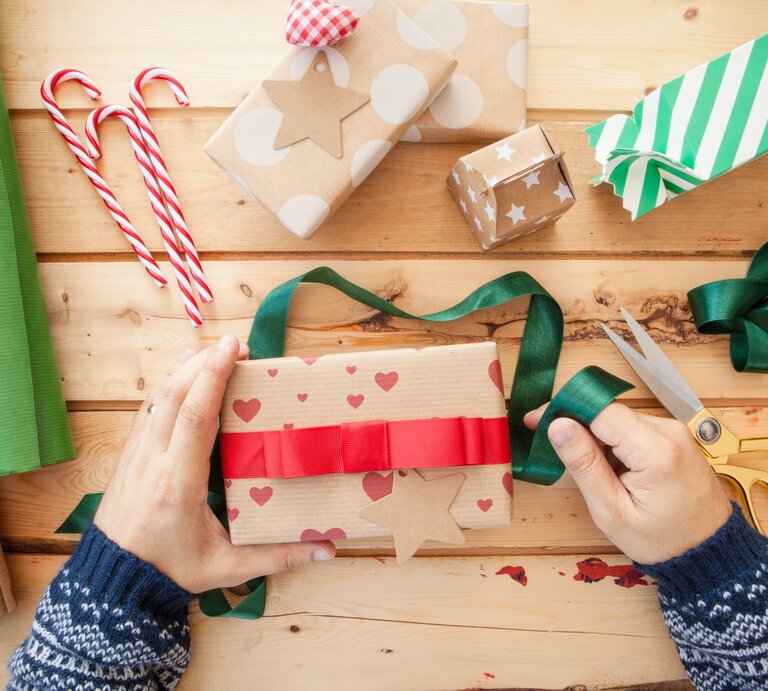 Zwei Hände in blauem Strickpulli verpacken ein Geschenk in Herzpapier mit rotem Band, umgeben von Geschenkpapier, Schleifen und Zuckerstangen. | © Adobe Stock/scerpica