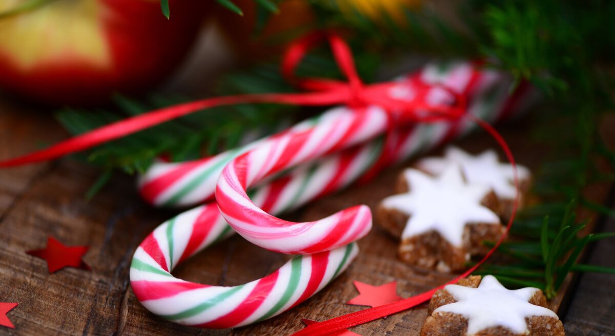 Zwei rot-weiß-grüne Zuckerstangen liegen auf Holz neben Zimtsternen, Sternkonfetti in Rot und grünen Tannenzweigen.