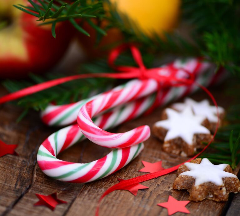 Zwei rot-weiß-grüne Zuckerstangen liegen auf Holz neben Zimtsternen, Sternkonfetti in Rot und grünen Tannenzweigen. | © Adobe Stock/Printemps
