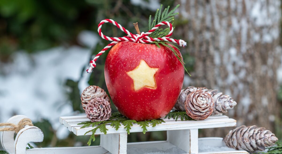 Ein roter Apfel mit ausgestochener Sternform und rot-weißer Kordel liegt auf einem weißen Schlitten, umgeben von Tannenzweigen und frostigen Zapfen.