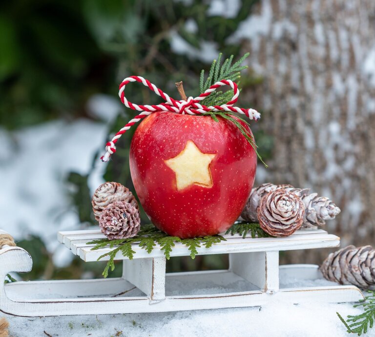 Ein roter Apfel mit ausgestochener Sternform und rot-weißer Kordel liegt auf einem weißen Schlitten, umgeben von Tannenzweigen und frostigen Zapfen. | © Adobe Stock/Natalia Greeske