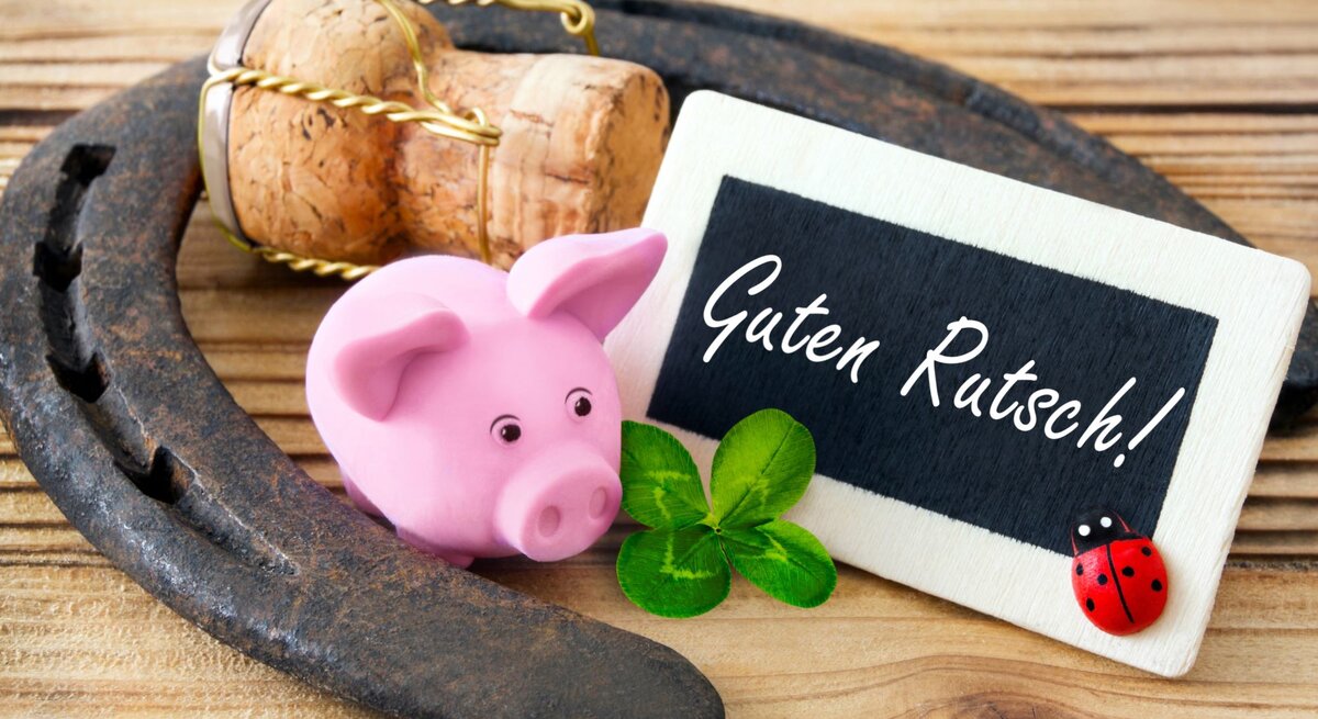 Ein rosafarbenes Glücksschwein, ein Kleeblatt, ein Hufeisen und eine Holztafel mit der Aufschrift „Guten Rutsch!“ liegen auf hellem Holz.