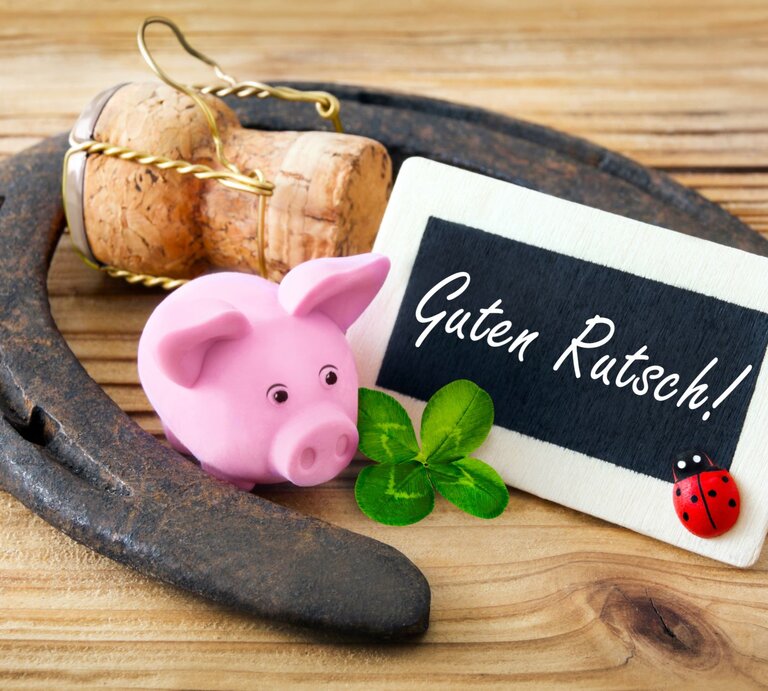 Ein rosafarbenes Glücksschwein, ein Kleeblatt, ein Hufeisen und eine Holztafel mit der Aufschrift „Guten Rutsch!“ liegen auf hellem Holz. | © Adobe Stock/PhotoSG