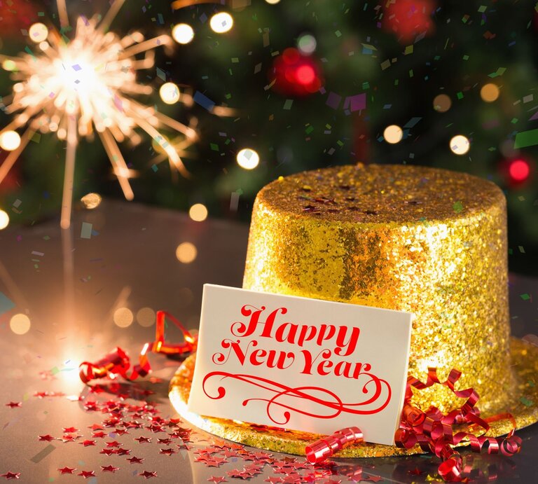 Ein glitzernder goldener Hut mit einer „Happy New Year“-Karte, roten Schleifen und Sternkonfetti vor einem leuchtenden Wunderkerzenhintergrund. | © Adobe Stock/WavebreakmediaMicro