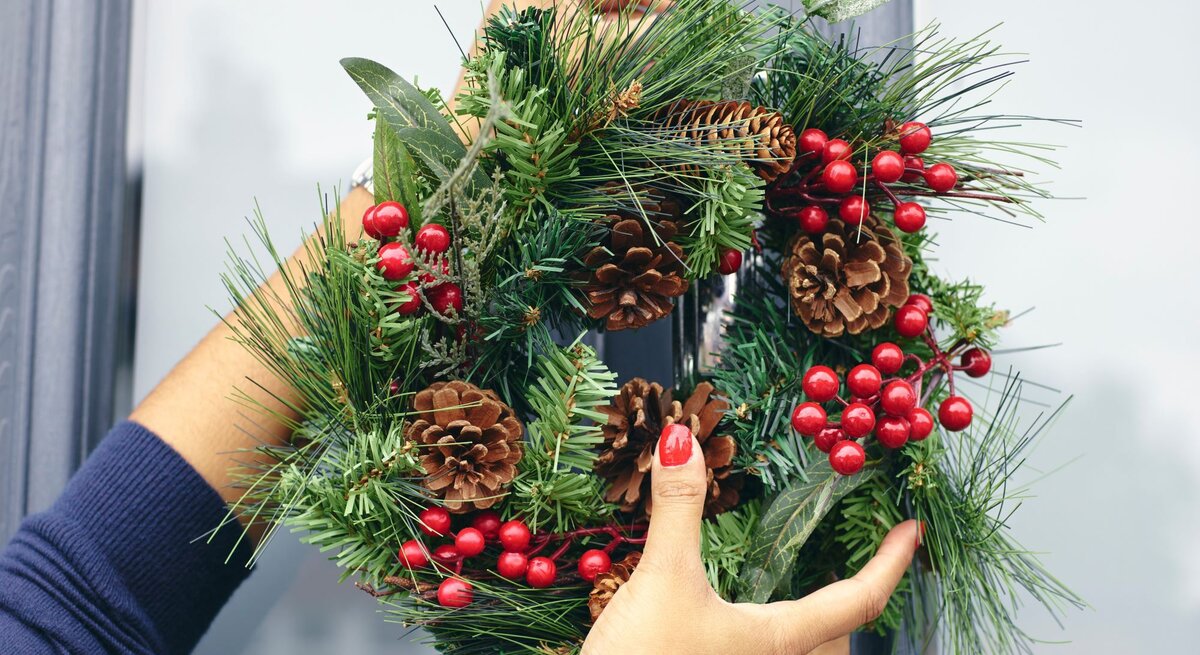 Hände einer Person mit rotem Nagellack hängen einen Weihnachtskranz aus Tannenzweigen, Zapfen und roten Beeren an eine blaue Tür.