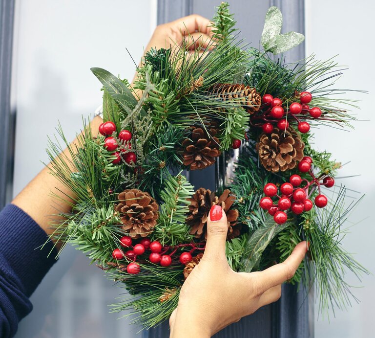 Hände einer Person mit rotem Nagellack hängen einen Weihnachtskranz aus Tannenzweigen, Zapfen und roten Beeren an eine blaue Tür. | © Adobe Stock/Cultura Creative