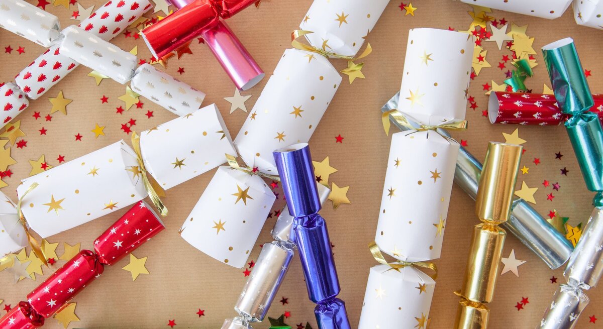 Bunte Silvester-Knallbonbons in Rot, Gold, Blau, Weiß und Grün liegen verstreut mit goldenen und roten Sternen auf braunem Papier.