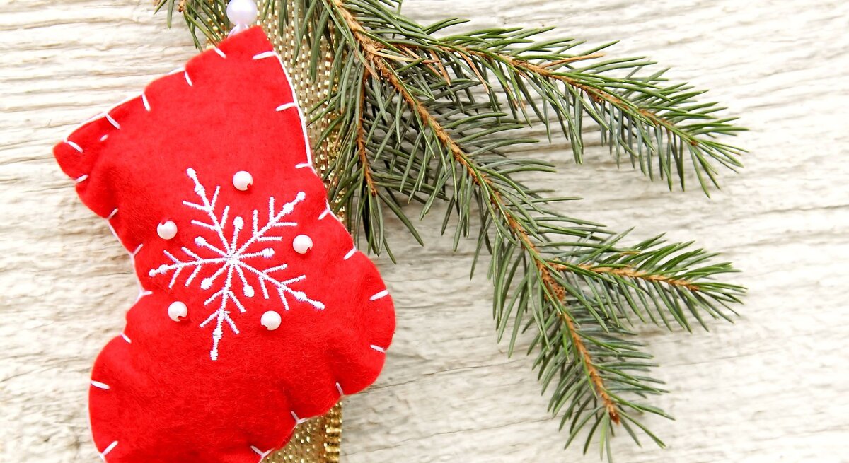 Eine rote Filz-Weihnachtssocke als Nikolausstiefel mit weißem Schneeflockenmotiv und Perlen hängt an einem Tannenzweig vor hellem Holz.