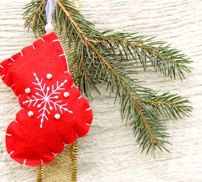 Eine rote Filz-Weihnachtssocke als Nikolausstiefel mit weißem Schneeflockenmotiv und Perlen hängt an einem Tannenzweig vor hellem Holz. | © Adobe Stock/Kathrin39