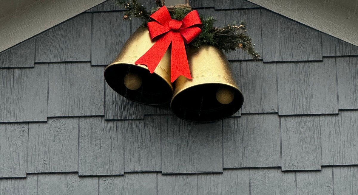 Zwei goldene Weihnachtsglocken mit roter Schleife und Tannenzweig hängen dekorativ an einer grau gestrichenen Hausfassade.