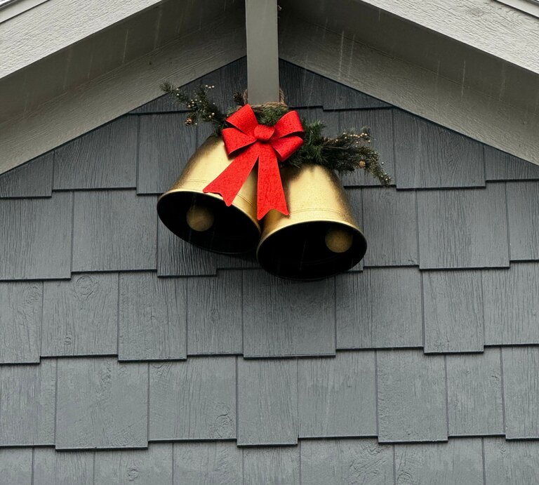 Zwei goldene Weihnachtsglocken mit roter Schleife und Tannenzweig hängen dekorativ an einer grau gestrichenen Hausfassade. | © Shutterstock/Ksuphil92
