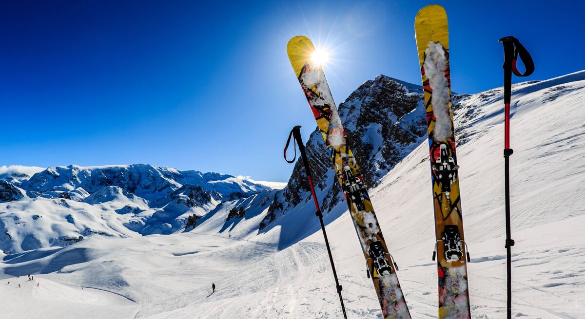 Zwei gelbe Ski und zwei Stöcke stehen im glitzernden Schnee vor einem sonnigen Bergpanorama mit tiefblauem Himmel.