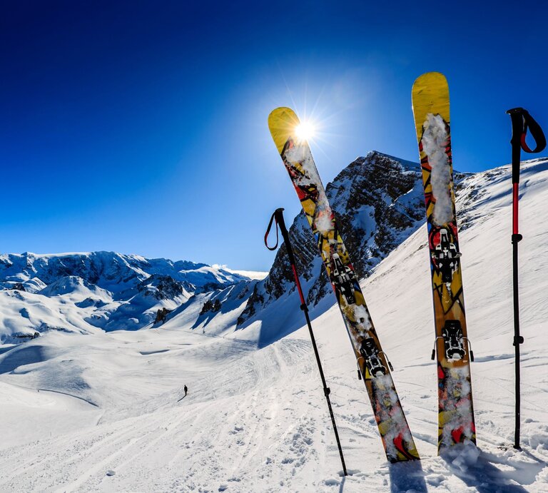 Zwei gelbe Ski und zwei Stöcke stehen im glitzernden Schnee vor einem sonnigen Bergpanorama mit tiefblauem Himmel. | © Adobe Stock/Gorilla