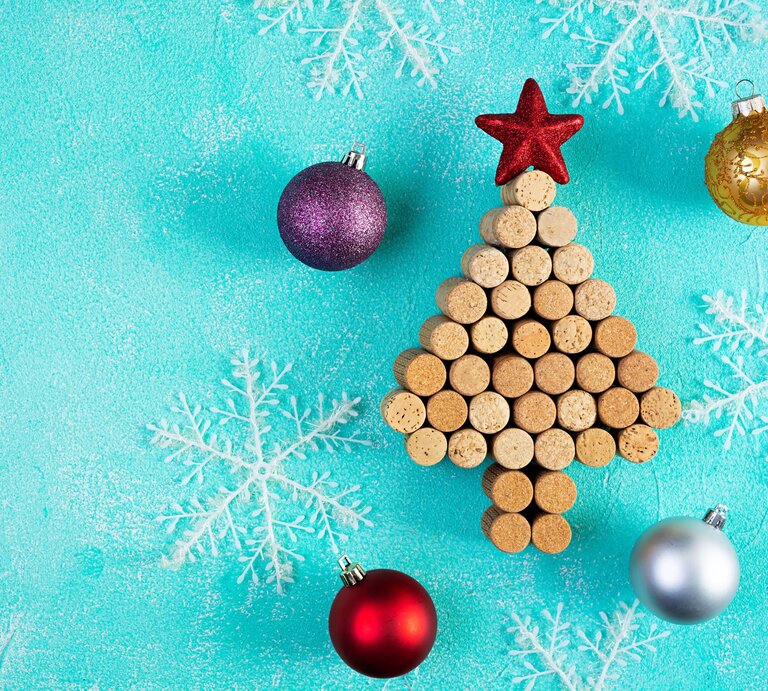 Aus Weinkorken gelegter Weihnachtsbaum mit rotem Stern, umgeben von Christbaumkugeln und weißen Schneeflocken auf türkisfarbenem Hintergrund. | © Adobe Stock/aeril