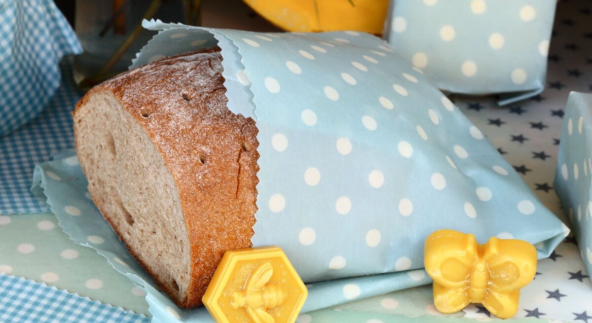 Ein halbes Brot in blau-weiß gepunktetem Bienenwachstuch, daneben gelbe Wachsfiguren in Schmetterlings- und Bienenform.