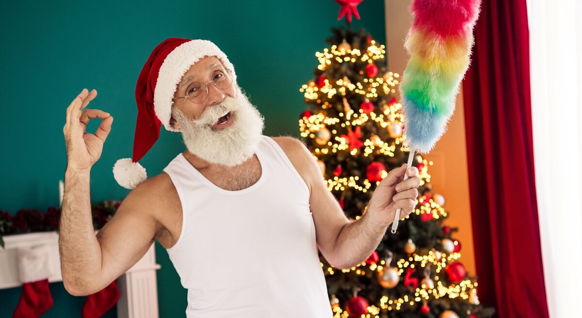 Basteln mit Staubwedel: Älterer Mann mit weißem Rauschebart und Santa-Mütze posiert lachend mit Staubwedel vor festlich geschmücktem Weihnachtsbaum.