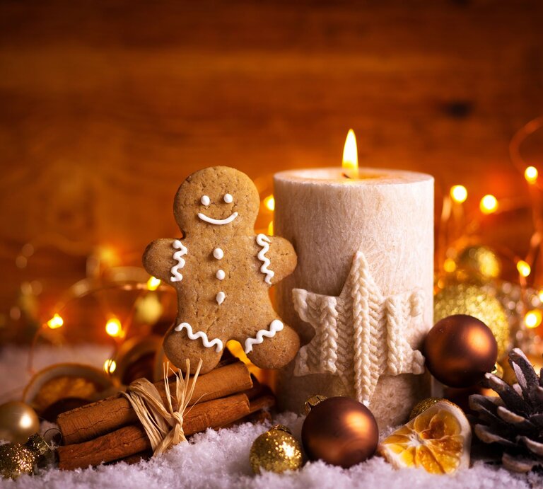 Lebkuchenmann neben einer brennenden Kerze mit Sternornament, umgeben von Zimtstangen, Tannenzapfen und weihnachtlichen Lichtern. | © Adobe Stock/Floydine