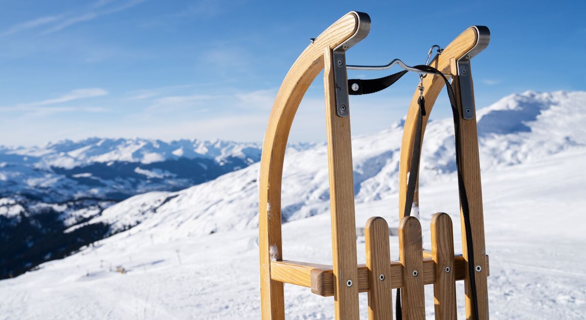 Ein hölzerner Schlitten steht auf schneebedecktem Bergplateau, im Hintergrund erstrecken sich sonnige Alpen unter klarem Himmel.