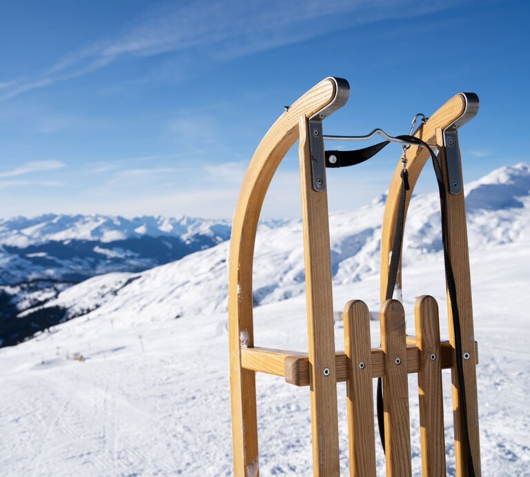 Ein hölzerner Schlitten steht auf schneebedecktem Bergplateau, im Hintergrund erstrecken sich sonnige Alpen unter klarem Himmel. | © Adobe Stock/Andrey Popov