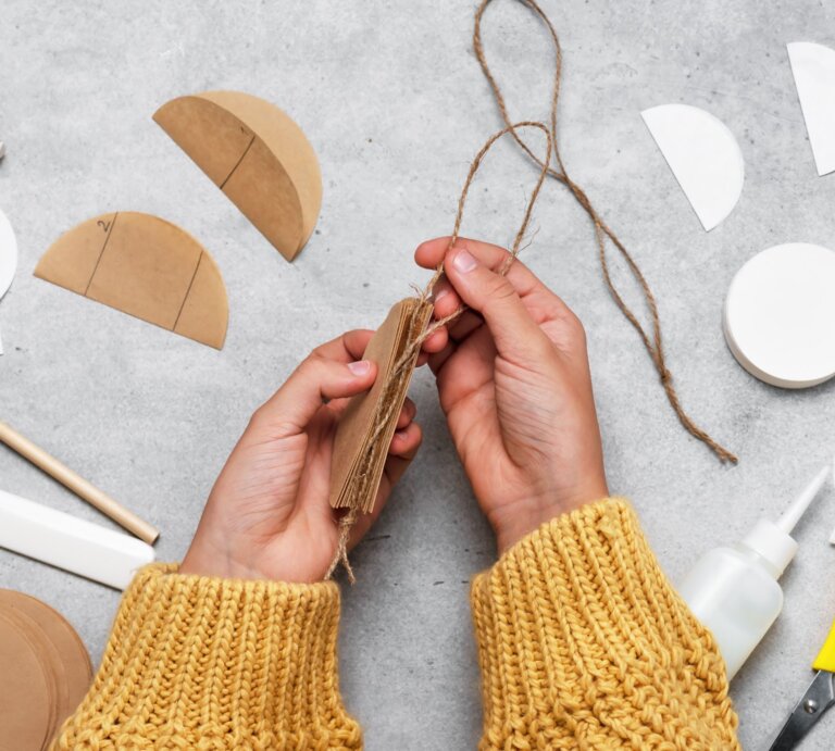 Hände in gelbem Strickpullover binden mit Juteschnur Pappscheiben zusammen, umgeben von Bastelmaterial, darunter Kaffeefilter, auf grauem Tisch. | © Adobe Stock/Oksana Schmidt