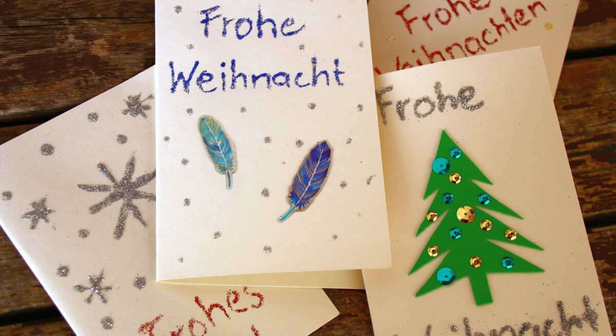 Handgemachte Weihnachtskarten mit Glitzersternen, Federdekoration und grünem Tannenbaum auf rustikalem Holztisch arrangiert.