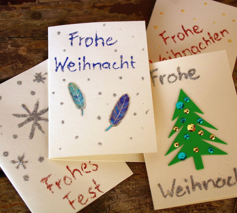 Handgemachte Weihnachtskarten mit Glitzersternen, Federdekoration und grünem Tannenbaum auf rustikalem Holztisch arrangiert. | © Adobe Stock/Christine