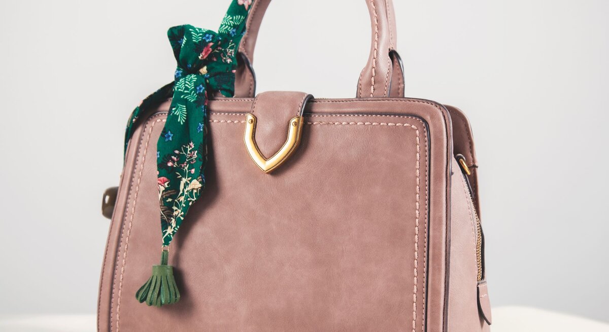 Beigefarbene Handtasche aus Leder mit goldener Verzierung und grünem, gemustertem Tuch an einem Henkel auf hellem Hintergrund.