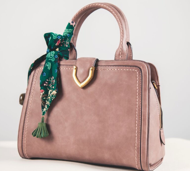 Beigefarbene Handtasche aus Leder mit goldener Verzierung und grünem, gemustertem Tuch an einem Henkel auf hellem Hintergrund. | © Adobe Stock/Alla