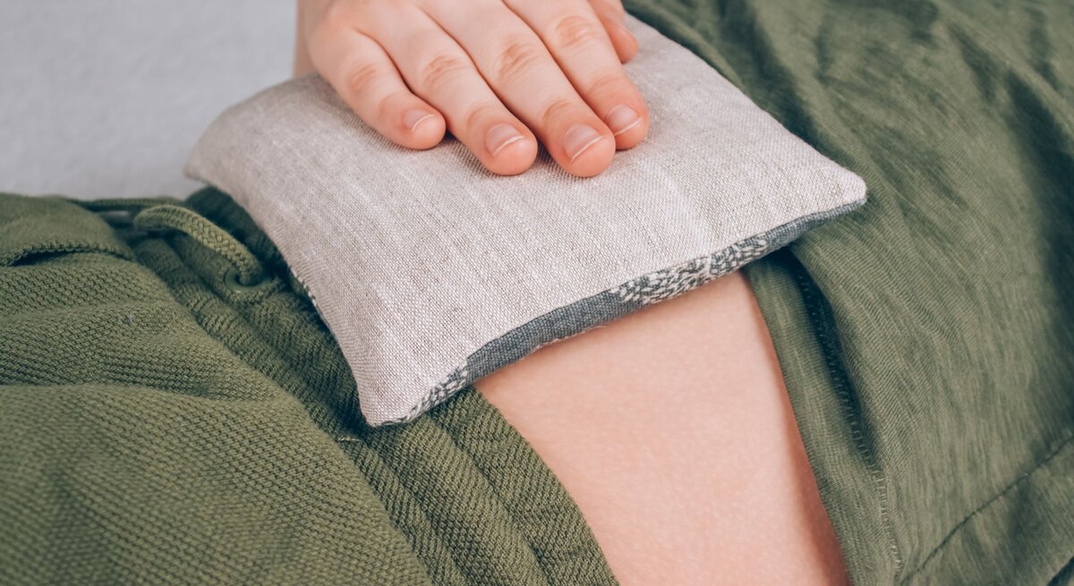 Eine Hand legt ein kleines Leinensäckchen auf den Bauch einer Person in grüner Kleidung zur Wärmeanwendung.