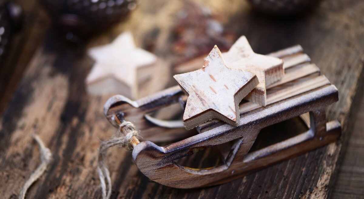 Kleine Holzschlitten-Dekoration mit zwei sternförmigen Anhängern auf rustikalem Holztisch, im Hintergrund unscharfe Weihnachtskugeln. Zwei weiße Holzsterne liegen auf einem kleinen Schlitten aus Holz, rustikaler Hintergrund mit Weihnachtsdeko. Detailaufnahme eines Mini-Holzschlittens mit Stern-Dekoration, rustikaler Holzuntergrund und verschwommene Ornamente im Hintergrund.