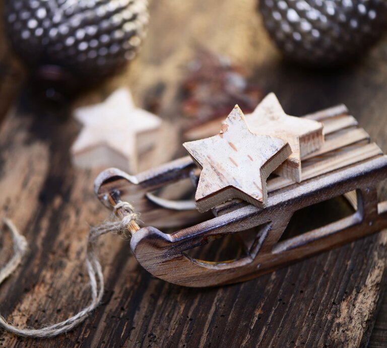 Kleine Holzschlitten-Dekoration mit zwei sternförmigen Anhängern auf rustikalem Holztisch, im Hintergrund unscharfe Weihnachtskugeln. Zwei weiße Holzsterne liegen auf einem kleinen Schlitten aus Holz, rustikaler Hintergrund mit Weihnachtsdeko. Detailaufnahme eines Mini-Holzschlittens mit Stern-Dekoration, rustikaler Holzuntergrund und verschwommene Ornamente im Hintergrund. | © Adobe Stock/photocrew