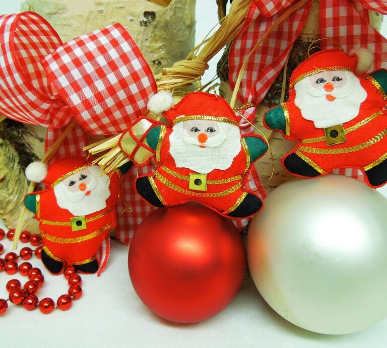 Drei Stoff-Weihnachtsmänner mit roten Anzügen hängen an einer karierten Schleife, daneben rote und silberne Kugeln. | © Adobe Stock/pipoint