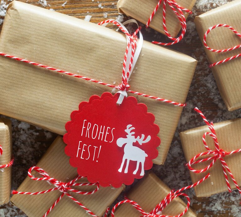Nahaufnahme von schlichten, braun verpackten Geschenken, verziert mit roten Kordeln und einem roten „Frohes Fest“-Anhänger. | © Adobe Stock/MissesJones