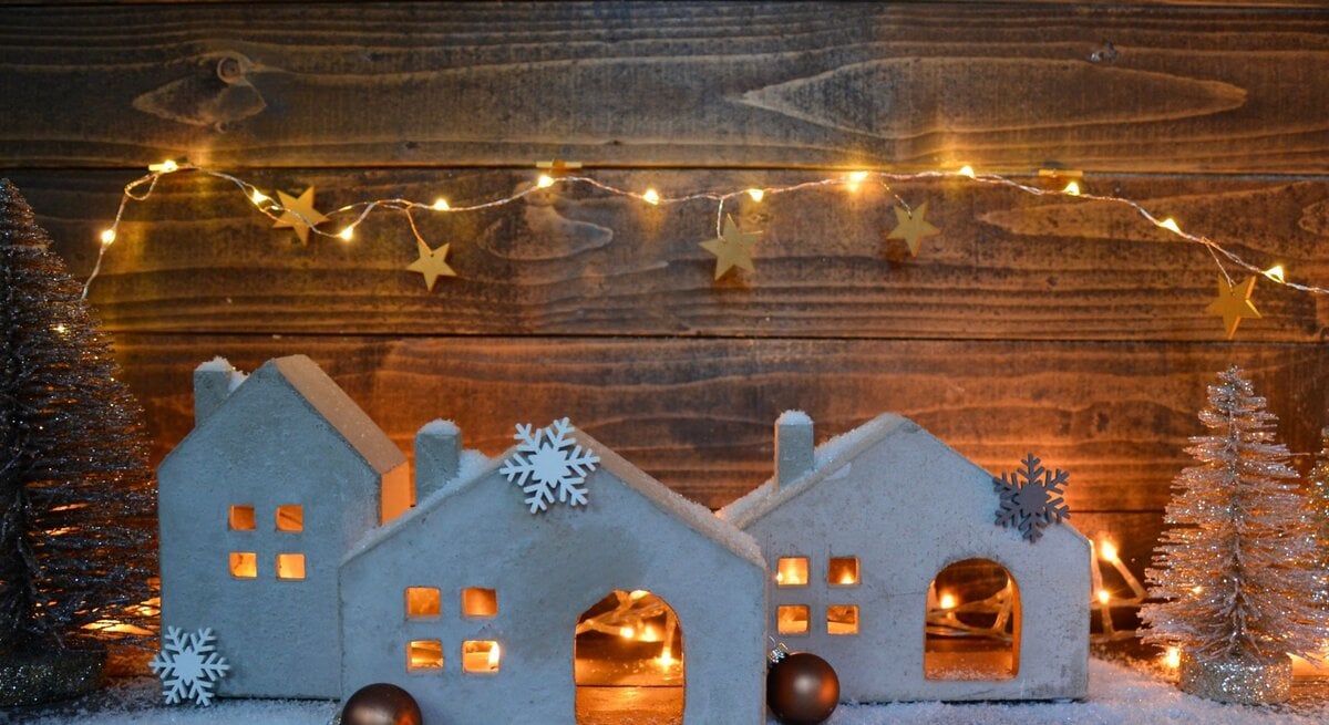 Kleine, weiße Häuser aus Ton mit Schneeflocken-Deko und Lichterkette stehen auf Kunstschnee vor einer Holzwand, umgeben von glitzernden Tannen.