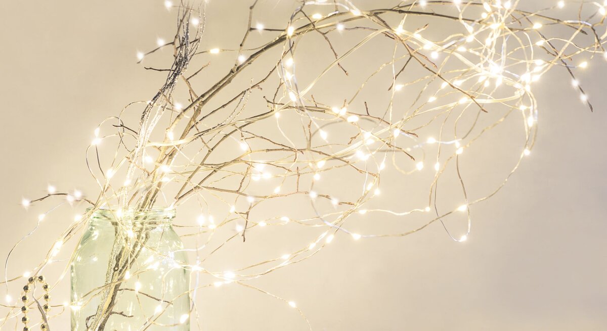 Ein bulliges glas mit Zweigen, die mit Lichterketten geschmückt sind, Hintergrund hell und die Lichterketten leuchten goldenweiss