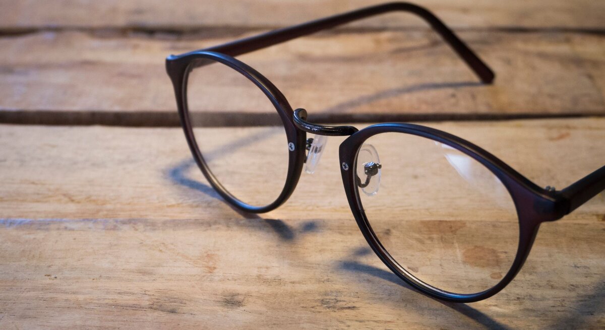Schwarze Brille mit runden Gläsern liegt auf einem rustikalen Holztisch in weichem, natürlichem Licht.