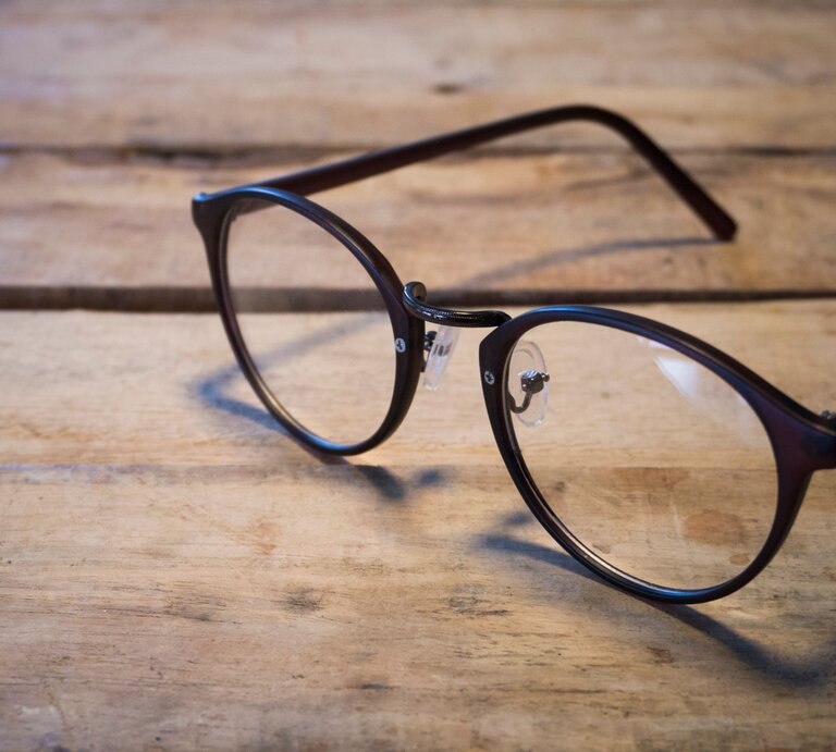 Schwarze Brille mit runden Gläsern liegt auf einem rustikalen Holztisch in weichem, natürlichem Licht. | © Adobe Stock/sugariie