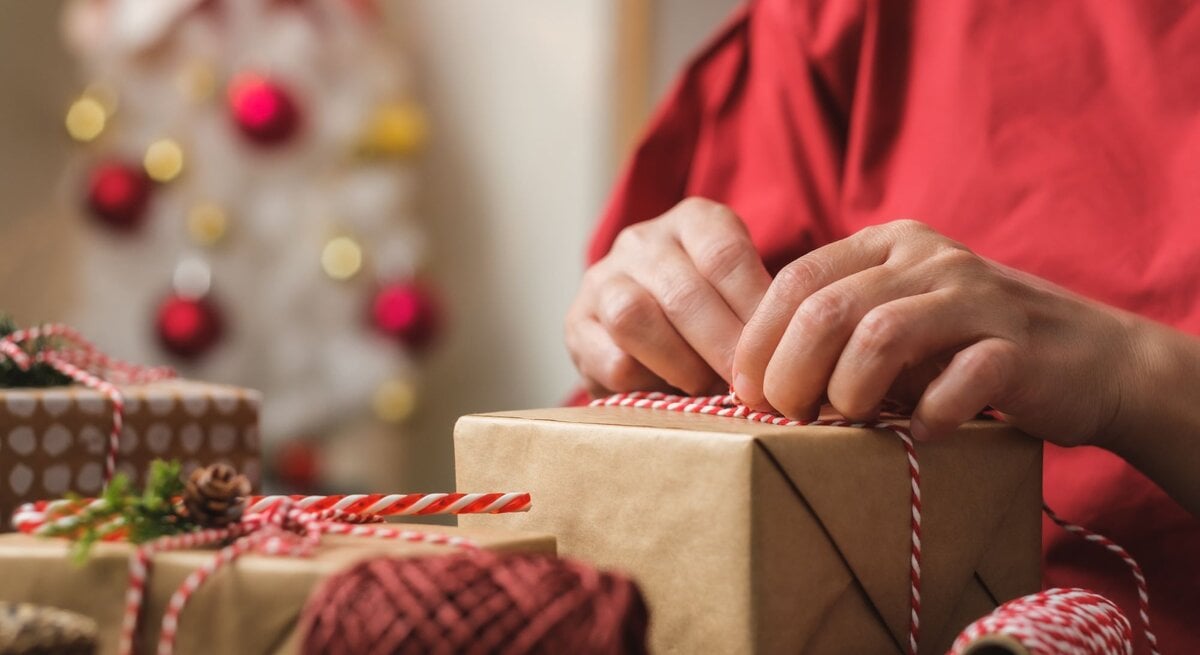 Eine Person bindet eine rot-weiße Schnur um ein Geschenk, im Hintergrund ein unscharfer Weihnachtsbaum mit Kugeln.