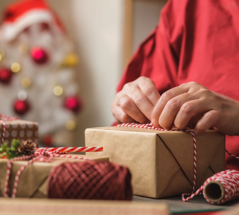 Eine Person bindet eine rot-weiße Schnur um ein Geschenk, im Hintergrund ein unscharfer Weihnachtsbaum mit Kugeln. | © Adobe Stock/weedezign