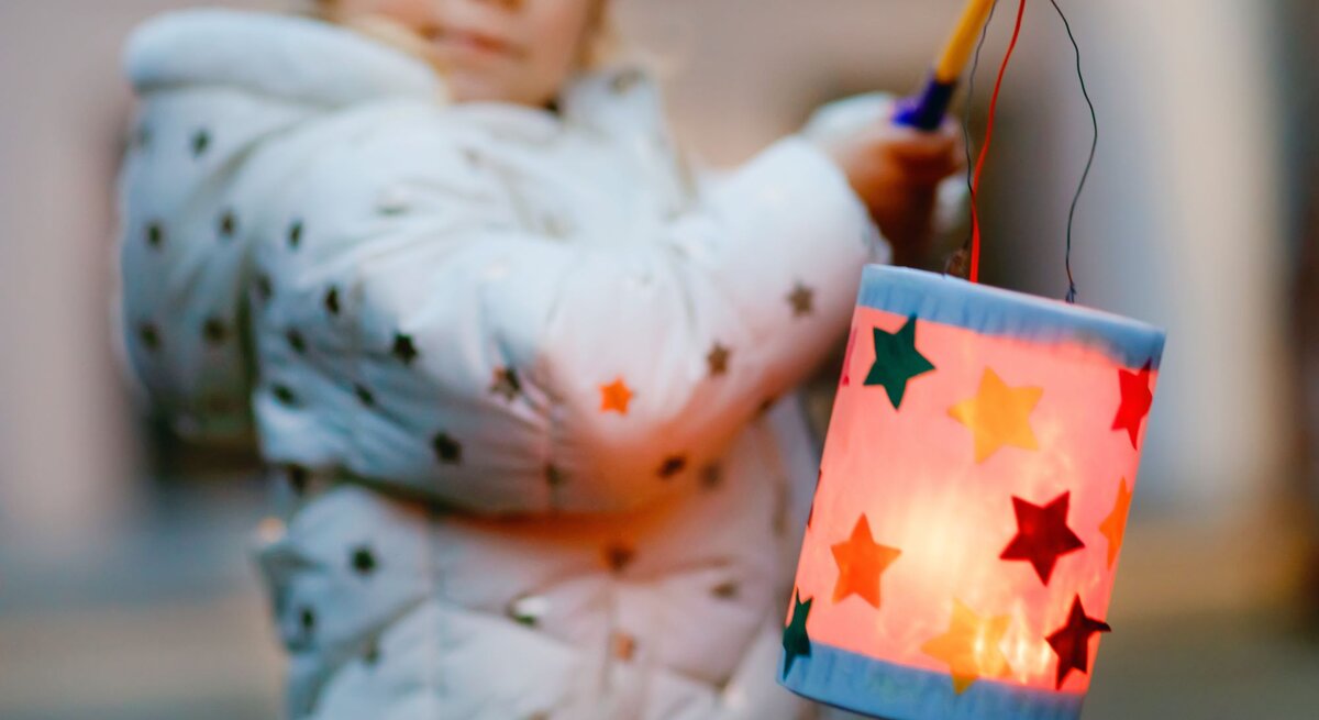 Ein kleines Kind in weißer Jacke mit Sternenmuster hält eine bunte Laterne mit leuchtenden Sternen vor verschwommenem Hintergrund.
