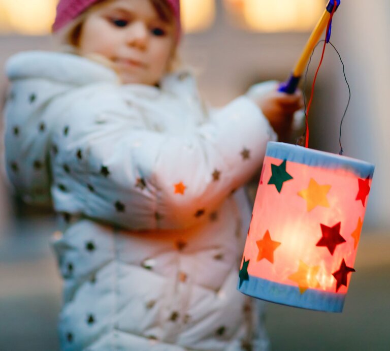 Ein kleines Kind in weißer Jacke mit Sternenmuster hält eine bunte Laterne mit leuchtenden Sternen vor verschwommenem Hintergrund. | © Adobe Stock/Irina Schmidt