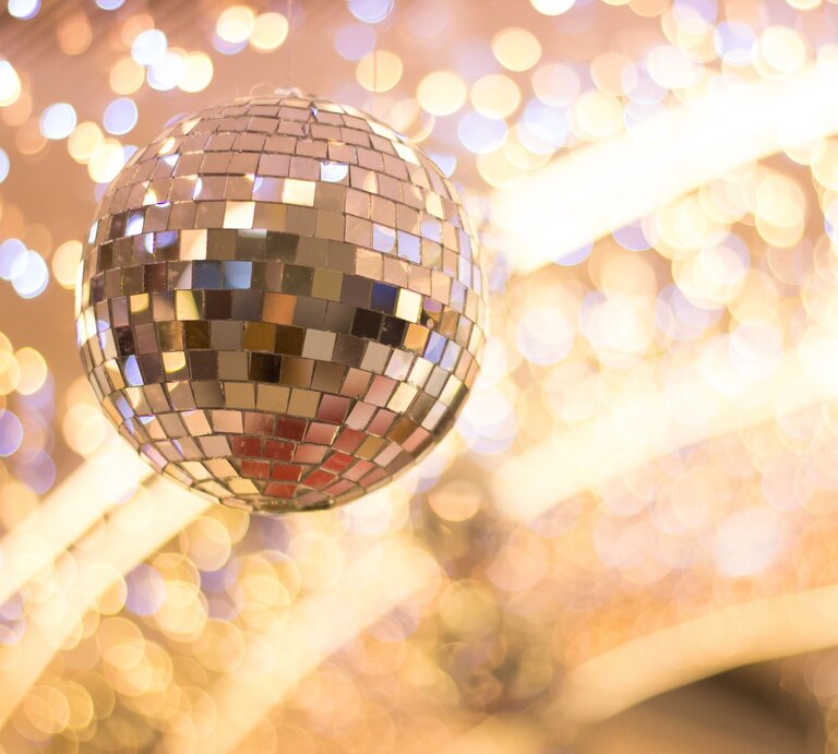 Nahaufnahme einer glitzernden Discokugel mit Spiegelmosaik, umgeben von warmen, verschwommenen Lichtreflexionen. | © Adobe Stock/Yuan