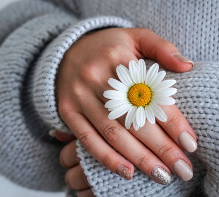 Zarte Frauenhände mit beiger und glitzernder Maniküre halten eine weiße Margerite auf grauem Strickpullover. | © Shutterstock/dutunectarie