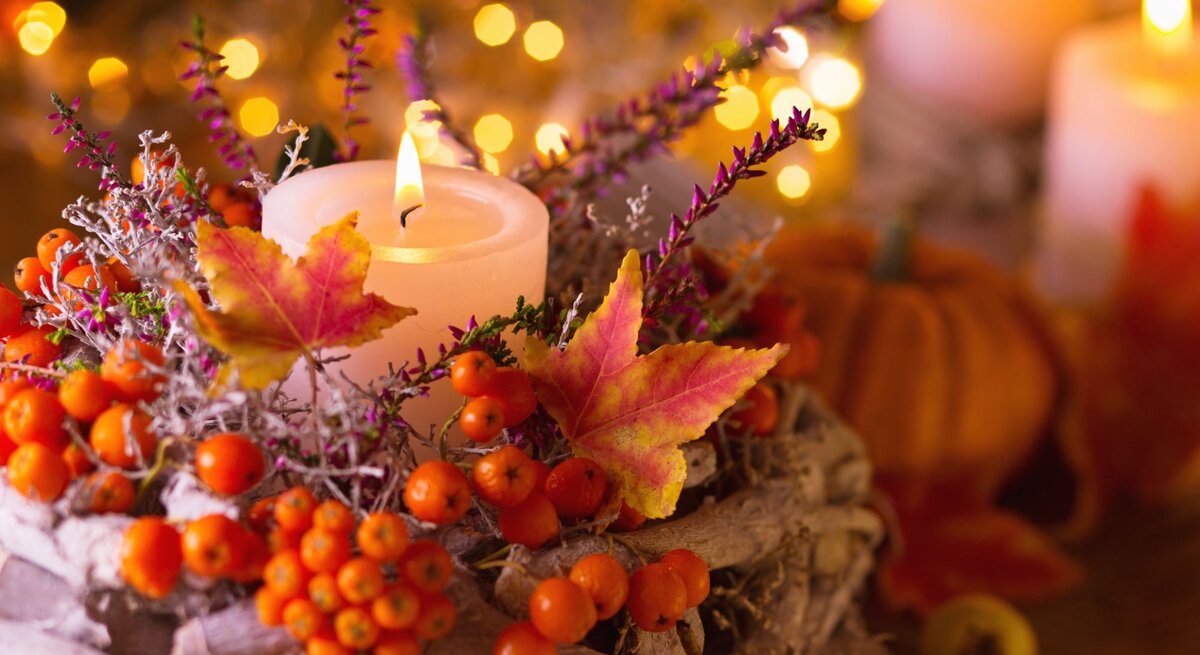 Ein brennendes Teelicht steht in einem herbstlichen Gesteck aus bunten Blättern, Beeren und Zweigen vor leuchtenden Lichterketten.