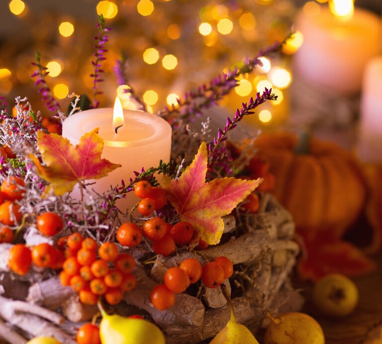Ein brennendes Teelicht steht in einem herbstlichen Gesteck aus bunten Blättern, Beeren und Zweigen vor leuchtenden Lichterketten. | © Adobe Stock/Floydine